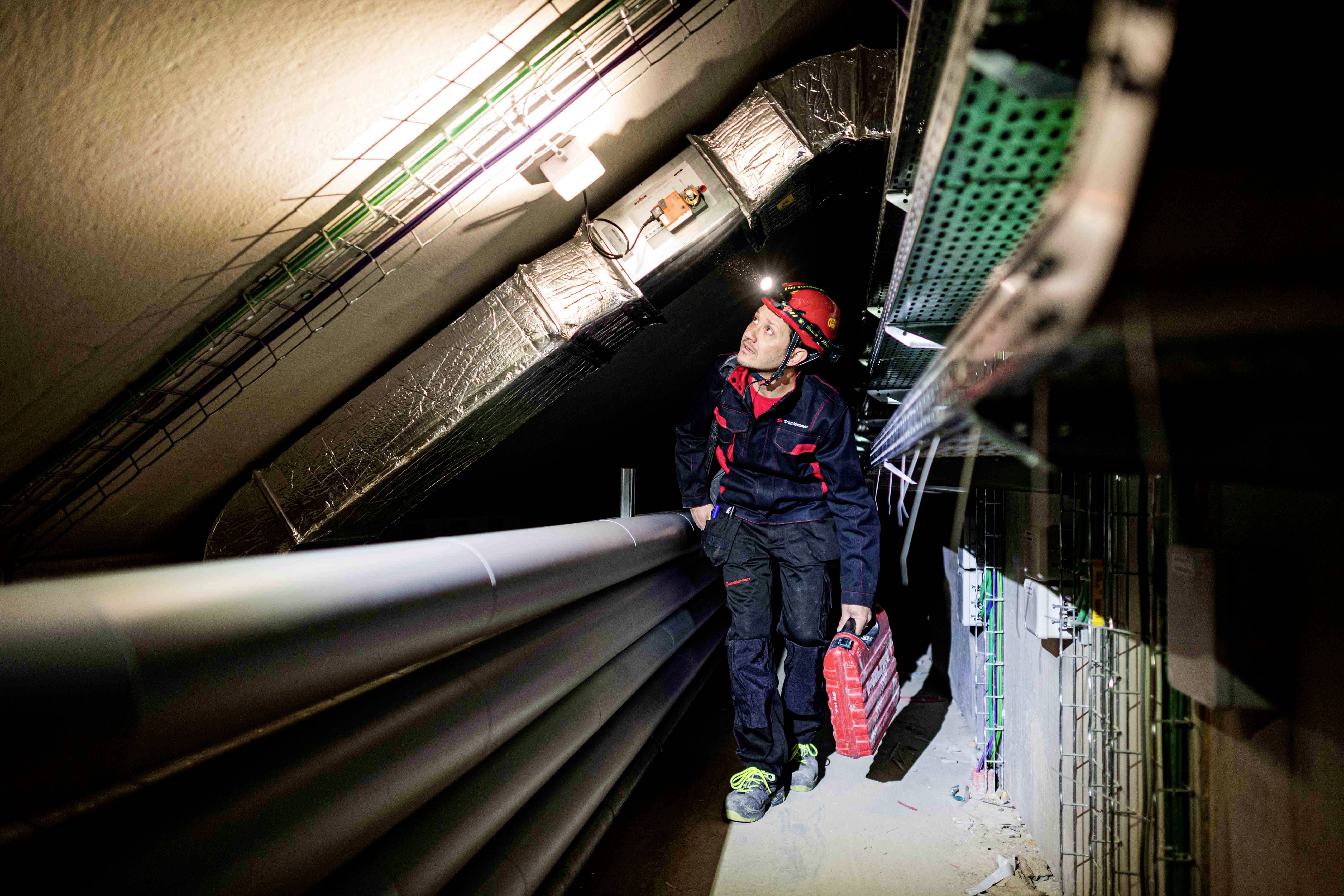 Elektryk w kasku i z walizką narzędziową kontroluje przewody w wąskim tunelu, oświetlonym przez czołówkę.