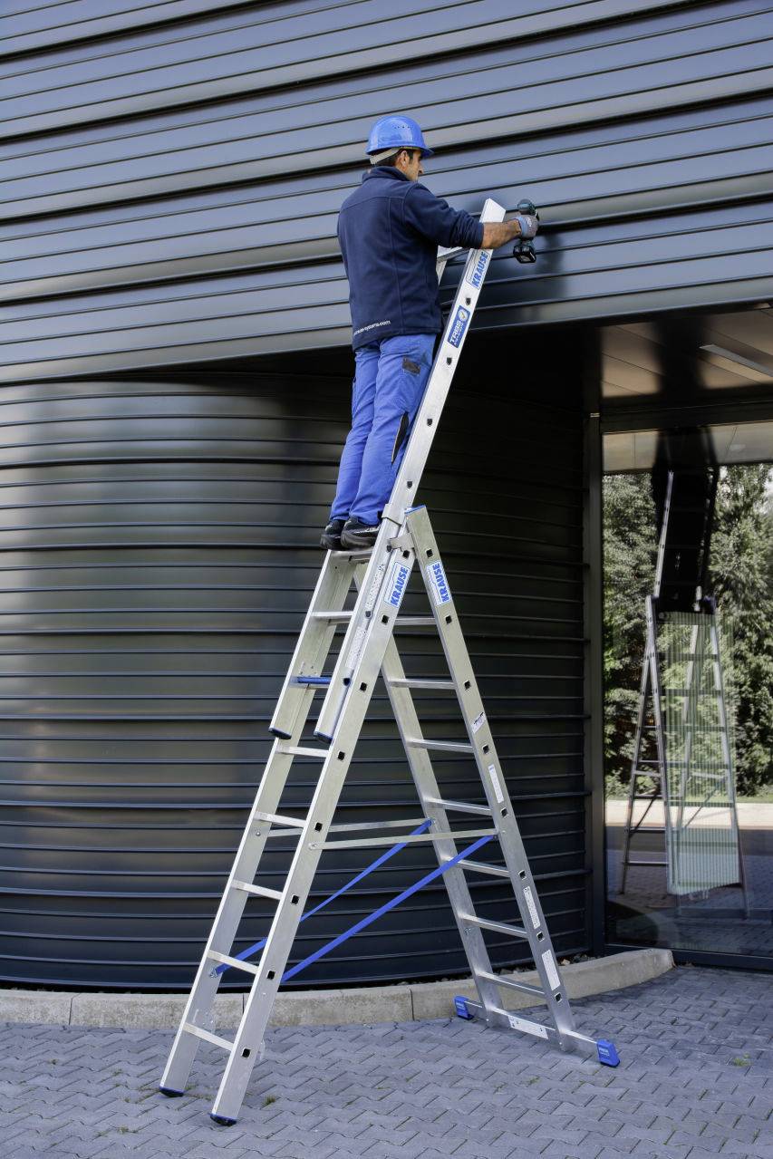 Osoba stoi na drabinie i mocuje coś do ściany budynku. Osoba jest ubrana w odzież ochronną i kask.