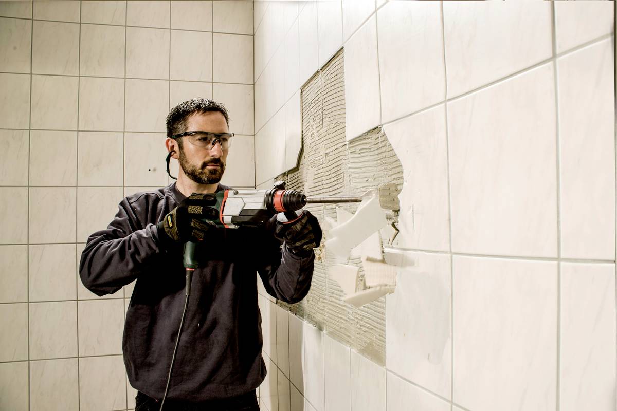 Mężczyzna w okularach ochronnych i rękawicach używa młotowiertarki, aby usunąć płytki ze ściany. Ściana betonowa jest częściowo widoczna.