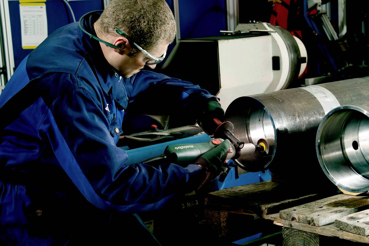 Robotnik w odzieży ochronnej szlifuje wewnętrzną stronę dużej metalowej rury elektronarzędziem w warsztacie.