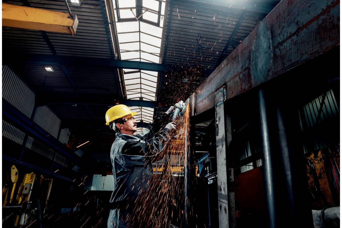 Robotnik w kasku ochronnym tnie metal elektryczną piłą w hali fabrycznej. Iskry rozbryzgują się we wszystkich kierunkach.