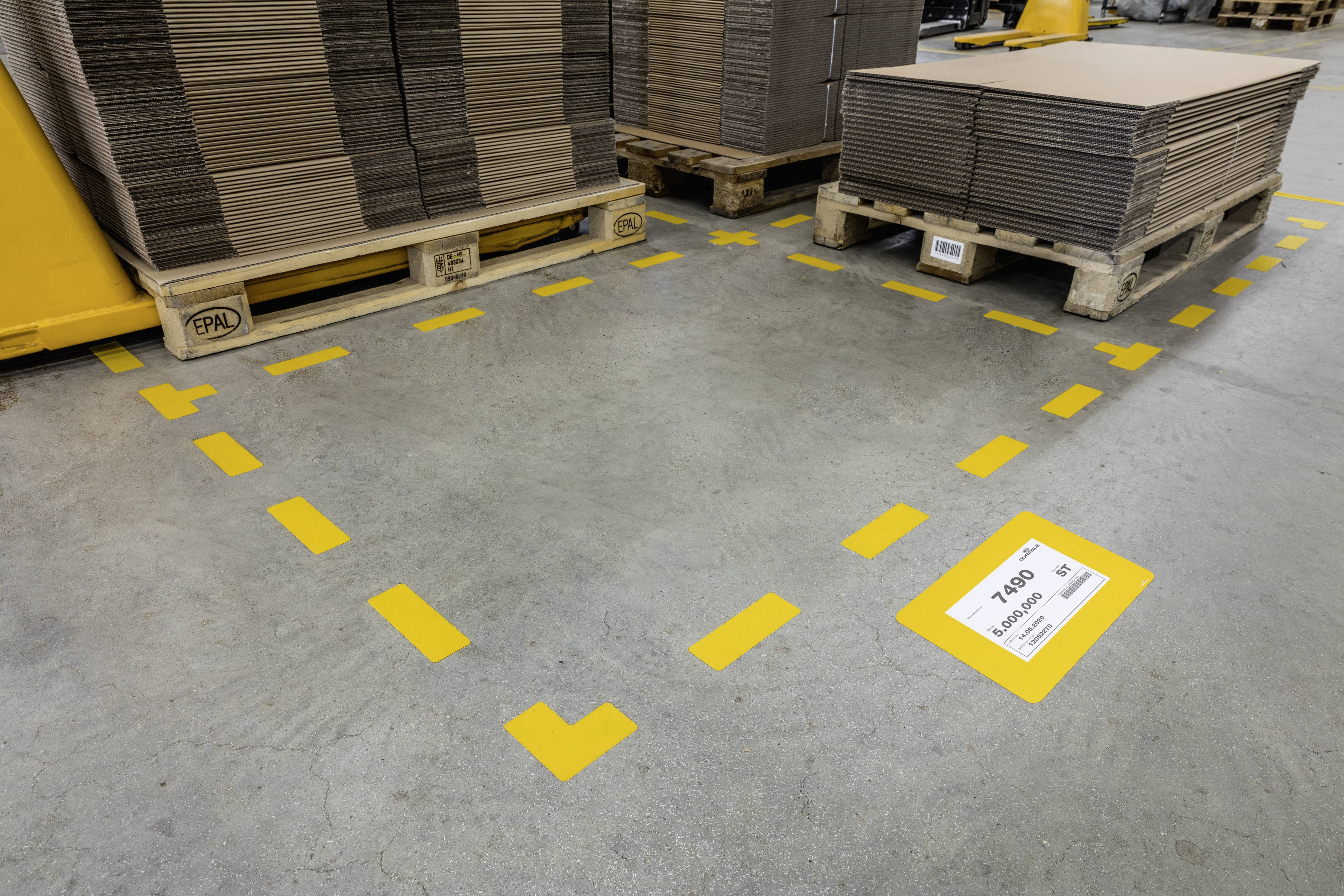 Kilka kartonowych pudeł ułożonych na paletach, bezpiecznie umieszczonych w oznaczonej żółtej strefie na podłodze magazynowej. Tabliczka wskazuje na zachowanie bezpiecznej odległości.