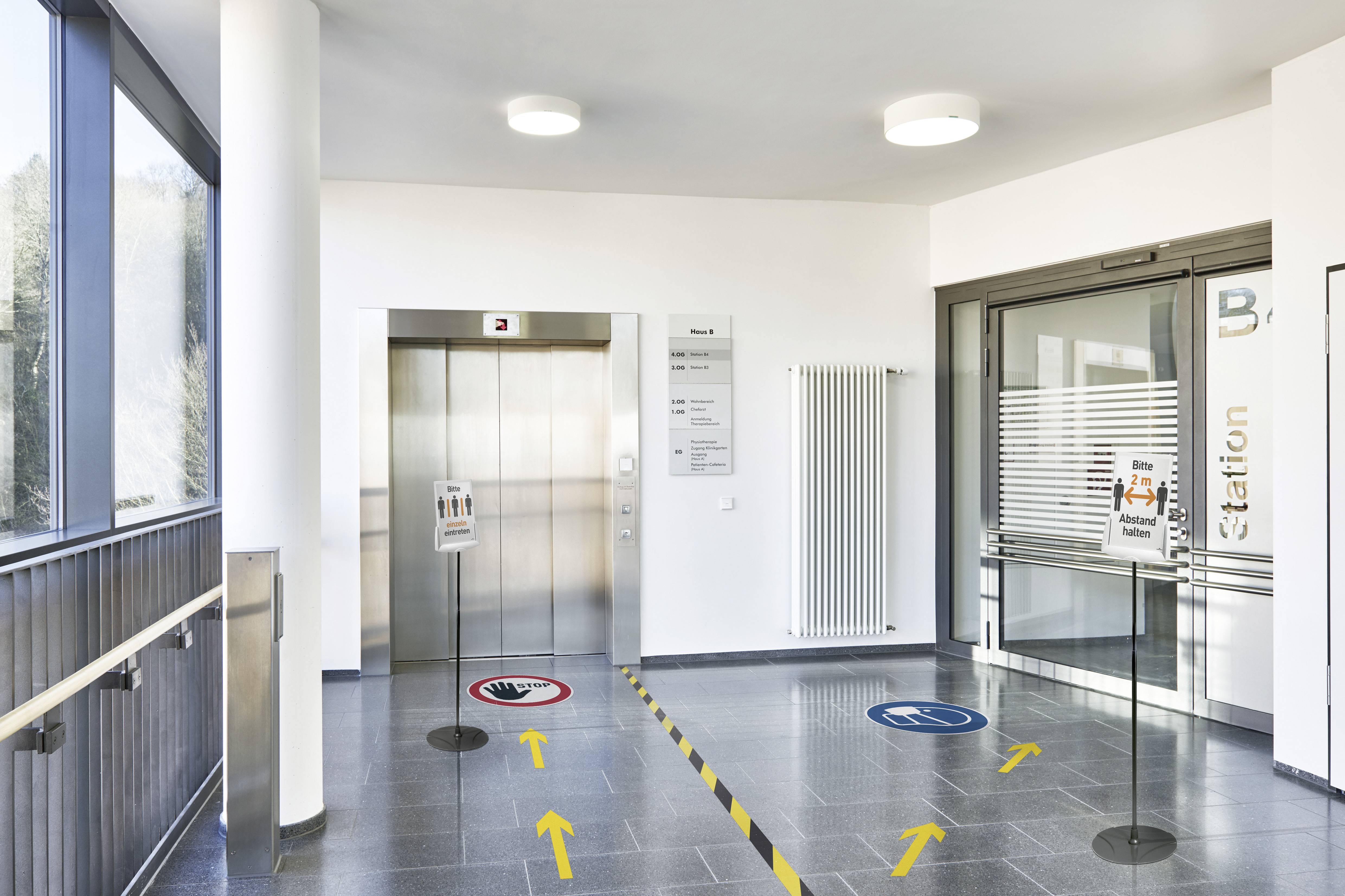 Wejście z windą. Oznaczenia na podłodze wskazują kierunki. Widoczne są tablice informacyjne dotyczące higieny i dostępności dla osób niepełnosprawnych.