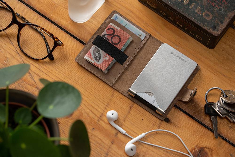 Portfel z banknotami i kartami leży obok okularów, słuchawek, kluczy i rośliny na drewnianym stole.