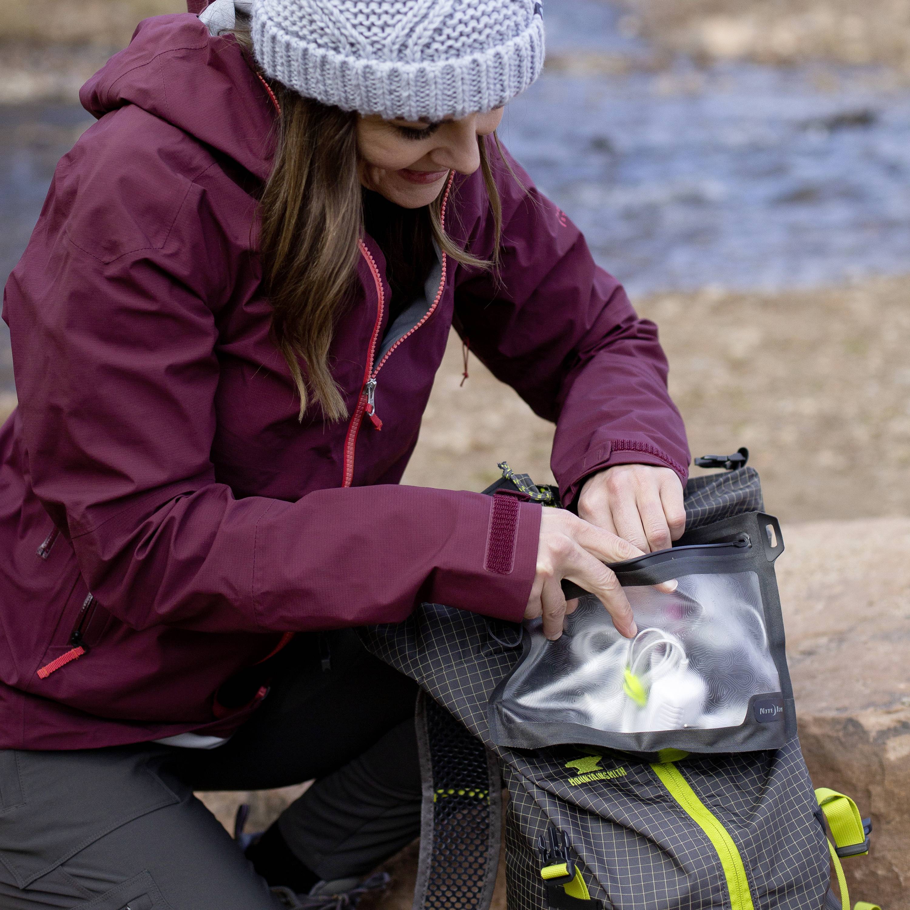 Osoba w czerwonej kurtce i szarej czapce pakuje przedmioty do szarego plecaka. W tle: skała i niewyraźny teren.