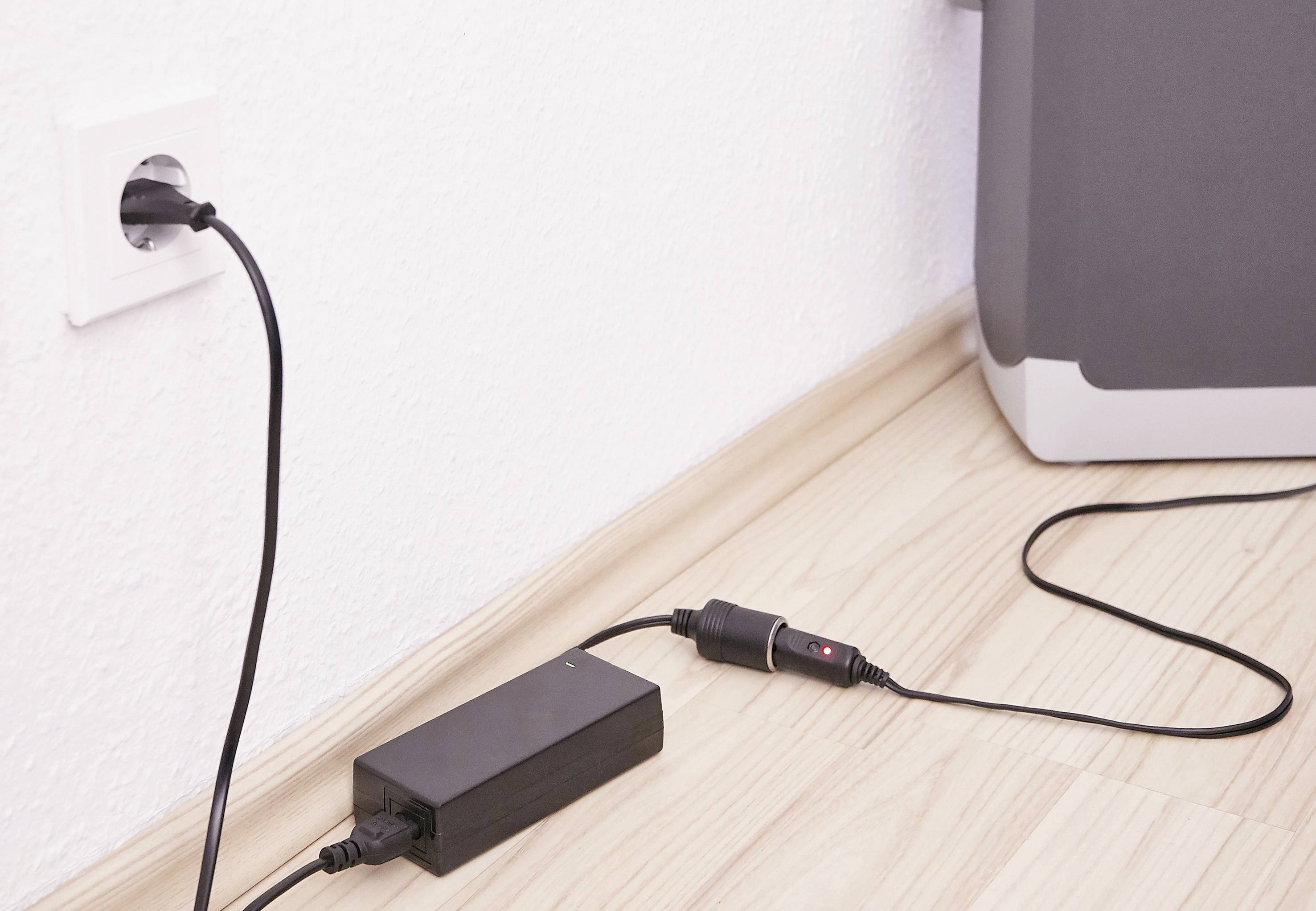 Czarna ładowarka jest podłączona do gniazdka elektrycznego. Kabel prowadzi do urządzenia leżącego na podłodze przed białą ścianą.