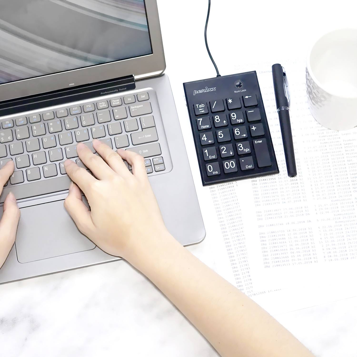 Osoba pisze na laptopie z klawiaturą numeryczną obok; na stole leży również kubek i długopis.