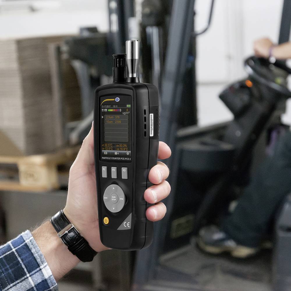 Osoba trzyma w ręku urządzenie do pomiaru cząstek. Na rozmytym tle widoczny jest wózek widłowy.