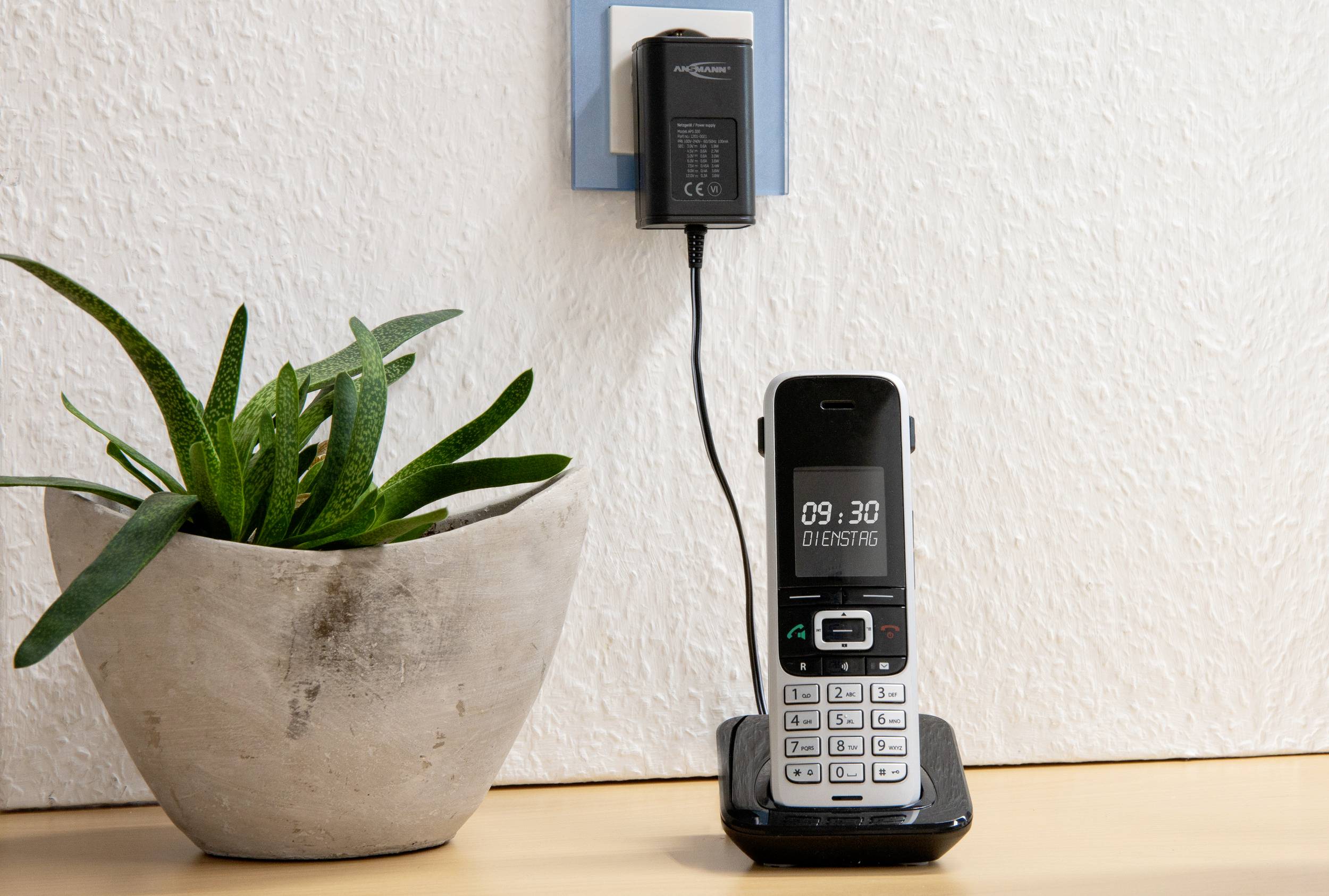 Bezprzewodowy telefon stacjonarny stoi na drewnianej półce obok zielonej rośliny w doniczce, telefon jest ładowany.