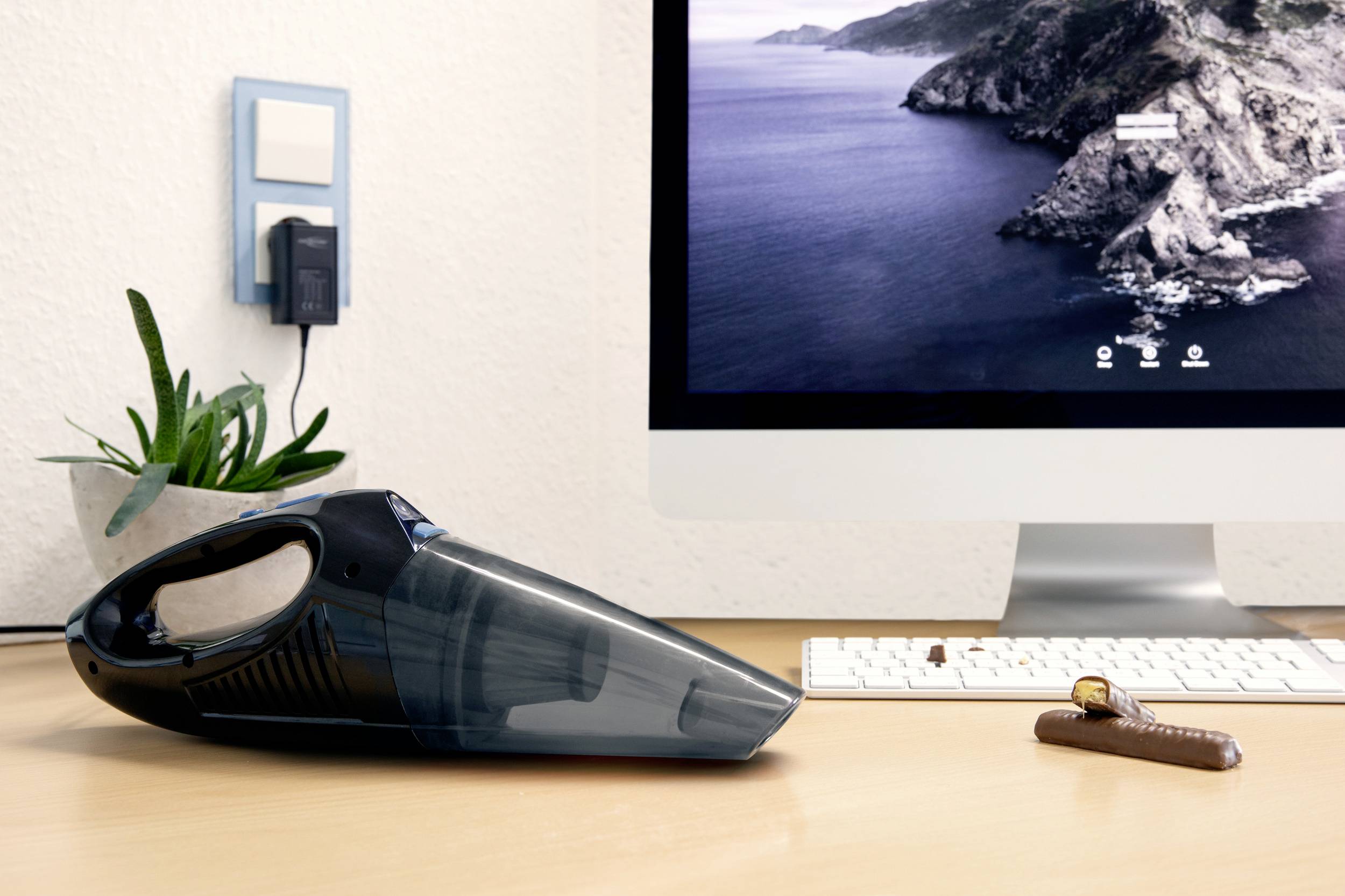 Poręczny odkurzacz leży na biurku przed ekranem komputera. Obok niego znajduje się batonik czekoladowy.