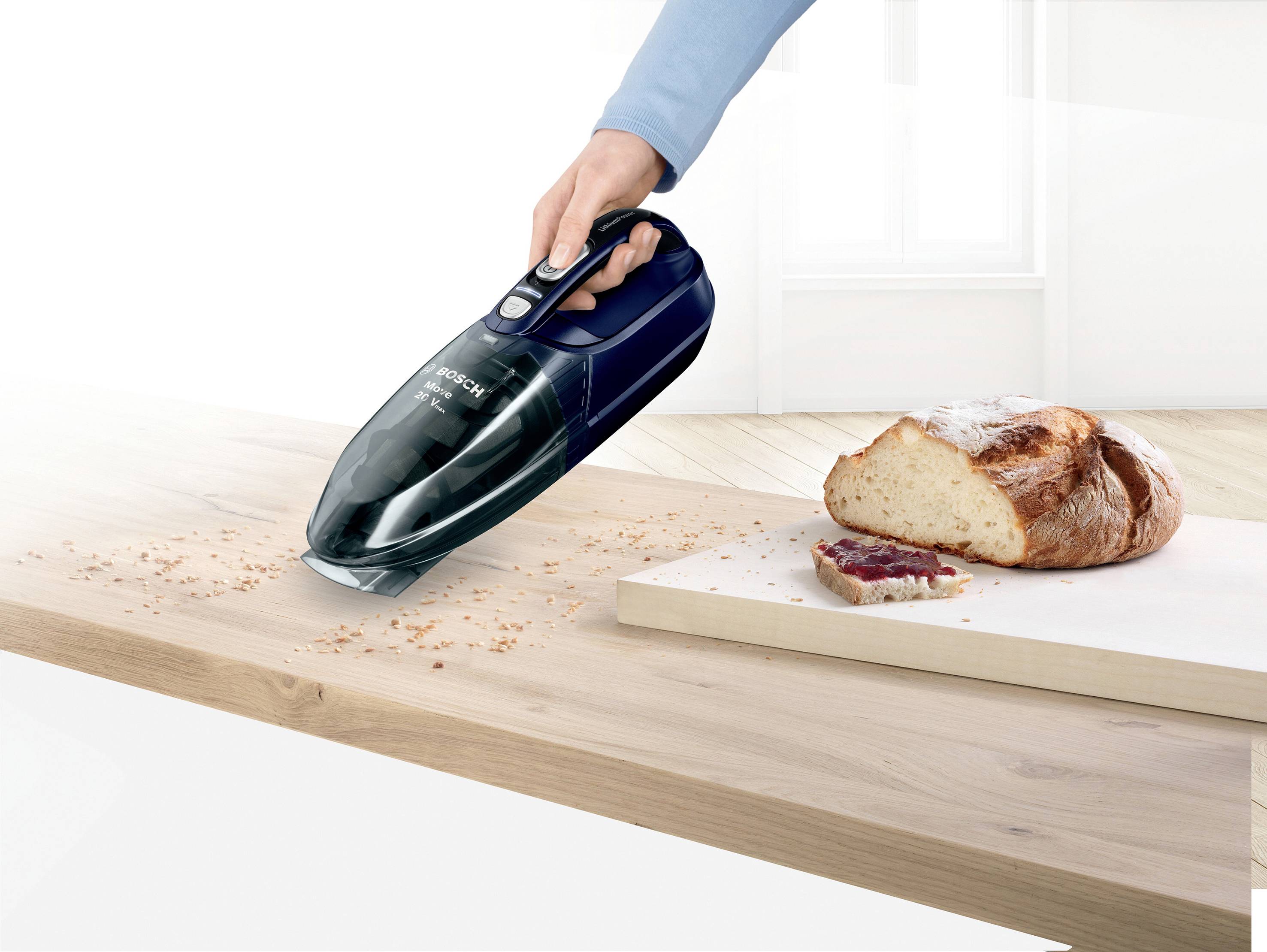 Jedna ręka trzyma ręczny odkurzacz, który usuwa okruchy ze stołu. Obok odkurzacza leży bochenek chleba z pokrojonym kawałkiem.