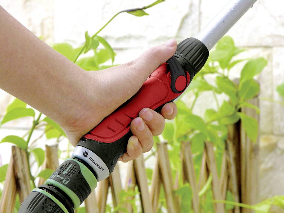 Ręka trzyma dyszę węża ogrodowego przed zielonymi roślinami i drewnianym płotem. Dysza jest częściowo czerwona z czarną rączką.