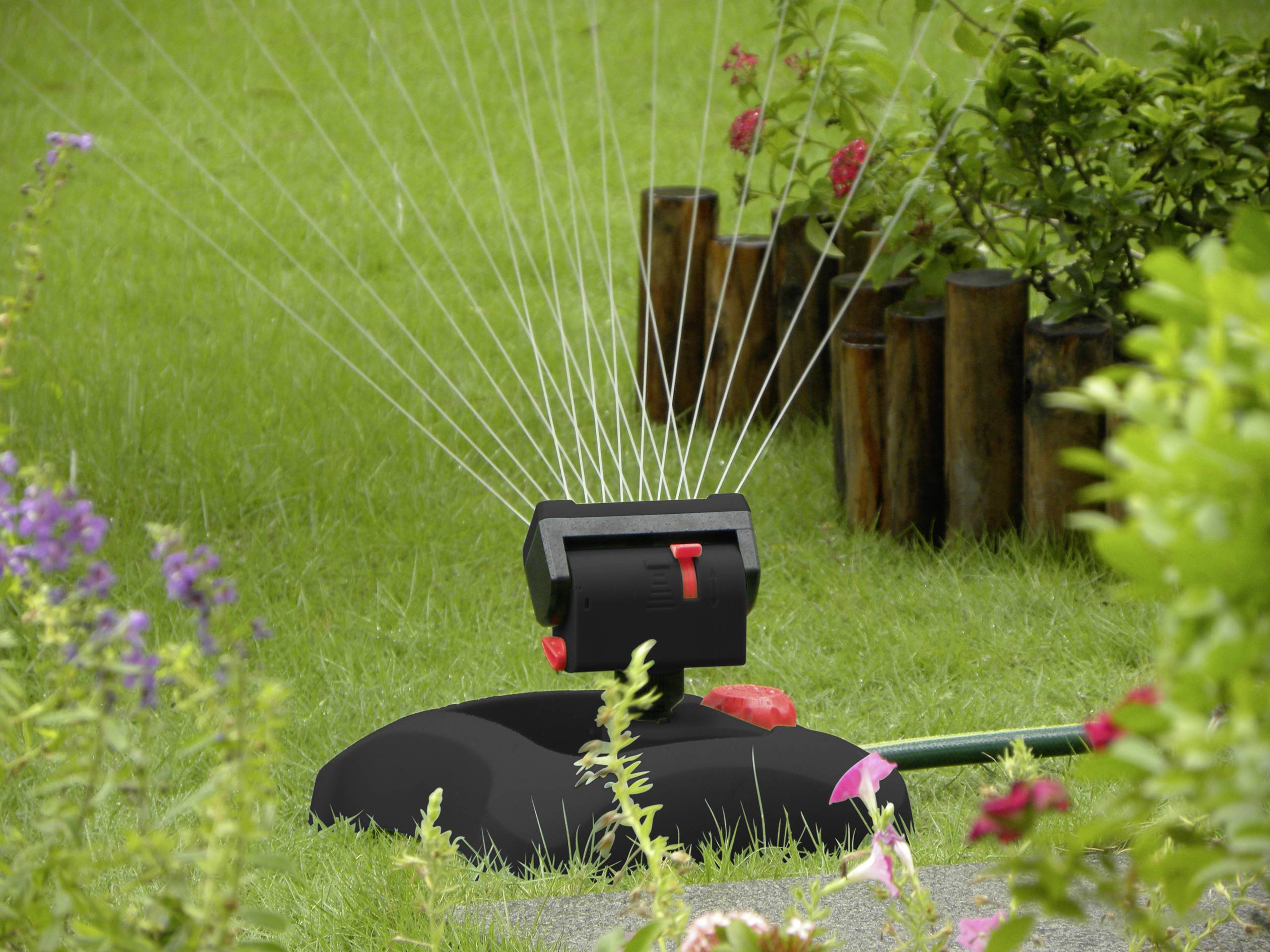 Zraszacz nawadnia zielony ogród; strugi wody tworzą równomierny półokrąg. Kwiaty i rośliny w tle.