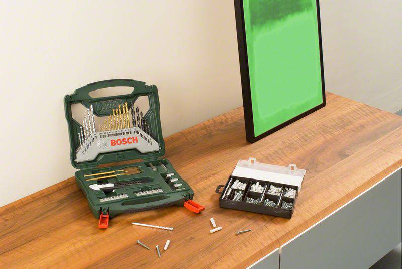 Zestaw wierteł z różnymi wiertłami i kołkami na drewnianym stole. Po prawej stronie na ścianie wisi obraz w zielonej ramce.
