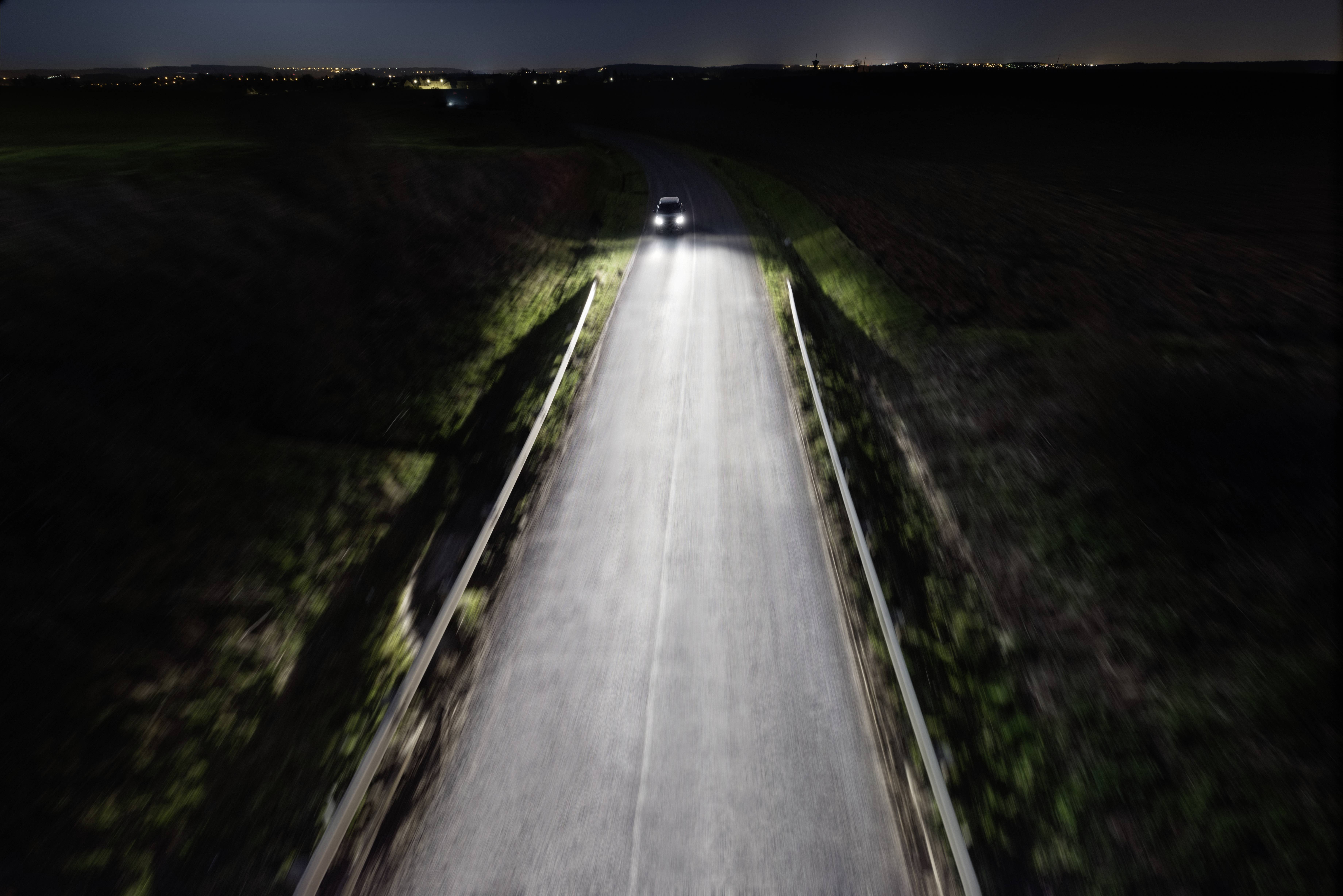 Samochód jedzie nocą po wąskiej, nieoświetlonej drodze. Reflektory oświetlają drogę. Otoczenie jest ciemne.