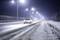 Samochód jedzie nocą po zaśnieżonej autostradzie, otoczony latarniami, które jasno oświetlają drogę.