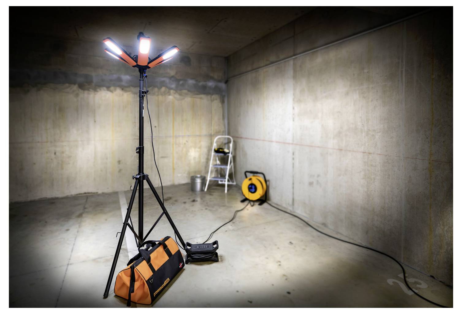 Przenośne lampy robocze na statywie oświetlają niedokończoną betonową piwnicę, a na podłodze leży drabina, kabel rozciągowy na bębnach i torba narzędziowa.