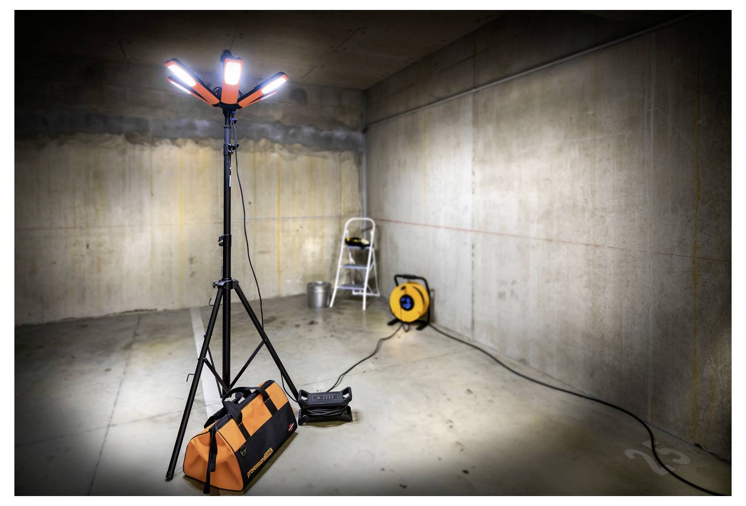 Lampa robocza na statywie i sprzęt w pustym pomieszczeniu z betonu, z drabiną i zwijaczem kabla sugerującymi przestrzeń w trakcie budowy lub przeglądu.