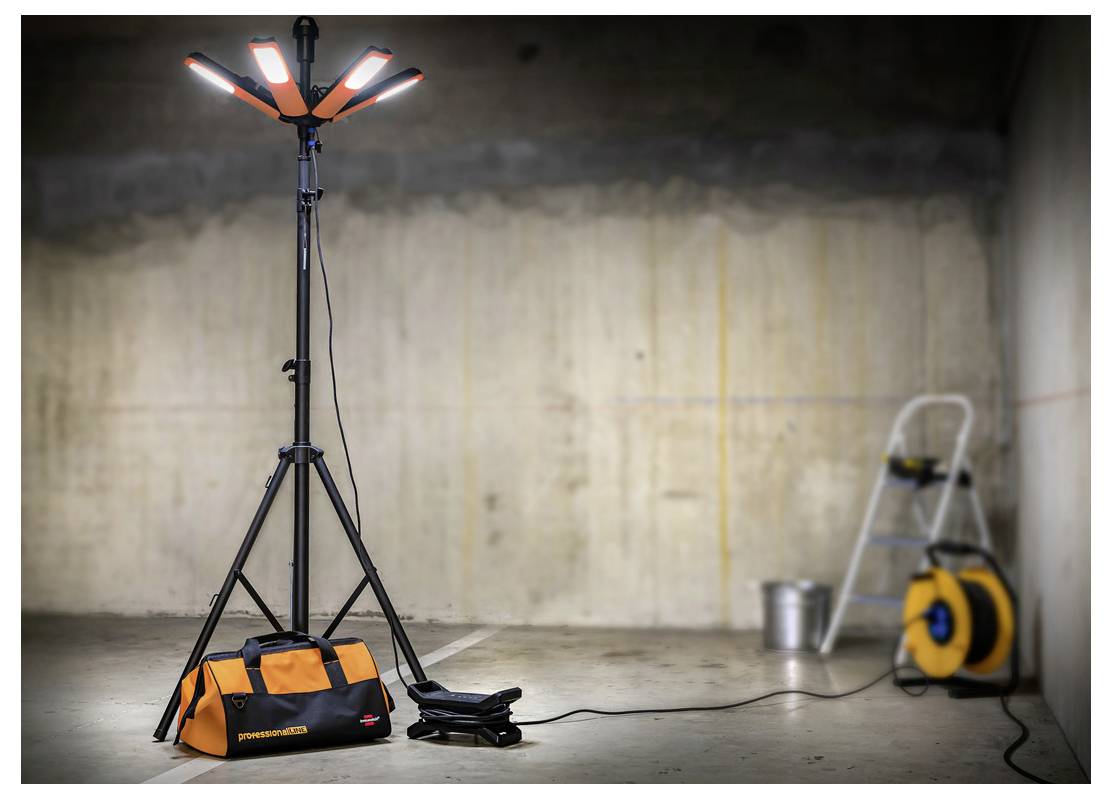 Przenośne światło robocze na statywie z pomarańczową torbą narzędziową i przedłużaczem w niedokończonym pokoju z drabiną i bębenem kabla w tle.