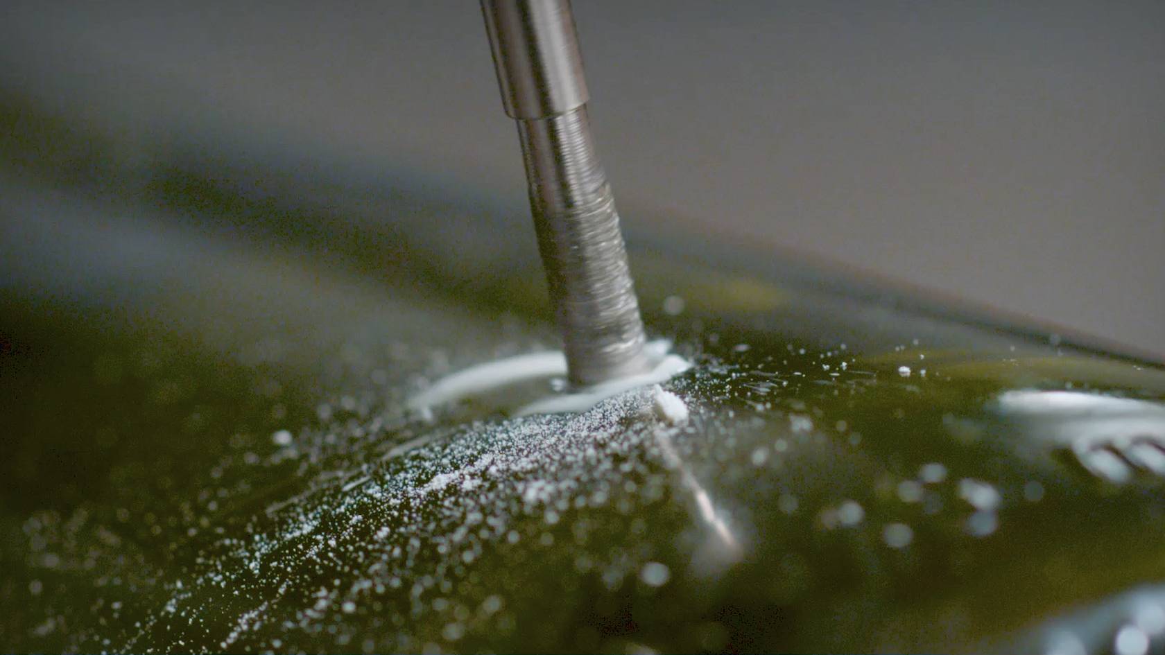 Zbliżenie wiertła ostrożnie wykonującego otwór w szklanej butelce, z wirującym wokół pyłem szklanym w centrum kadru.