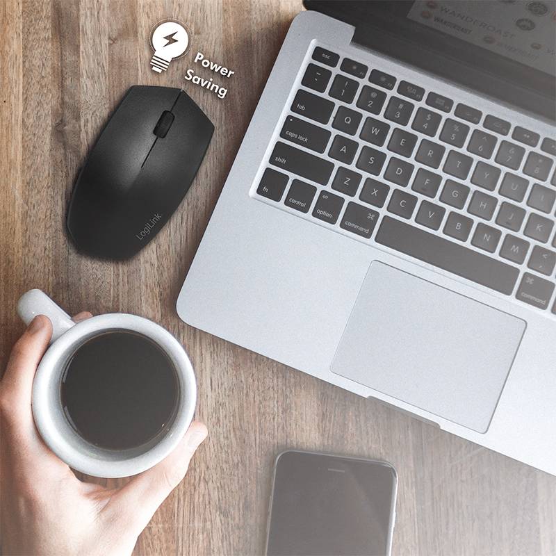 Laptop na drewnianej powierzchni, obok bezprzewodowa mysz z symbolem oszczędzania energii, osoba trzyma kubek kawy. Smartfon leży na stole.