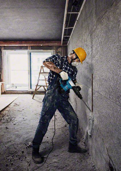Robotnik w kasku ochronnym i masce używa wiertarki przy betonowej ścianie na terenie budowy. W tle stoi drabina.