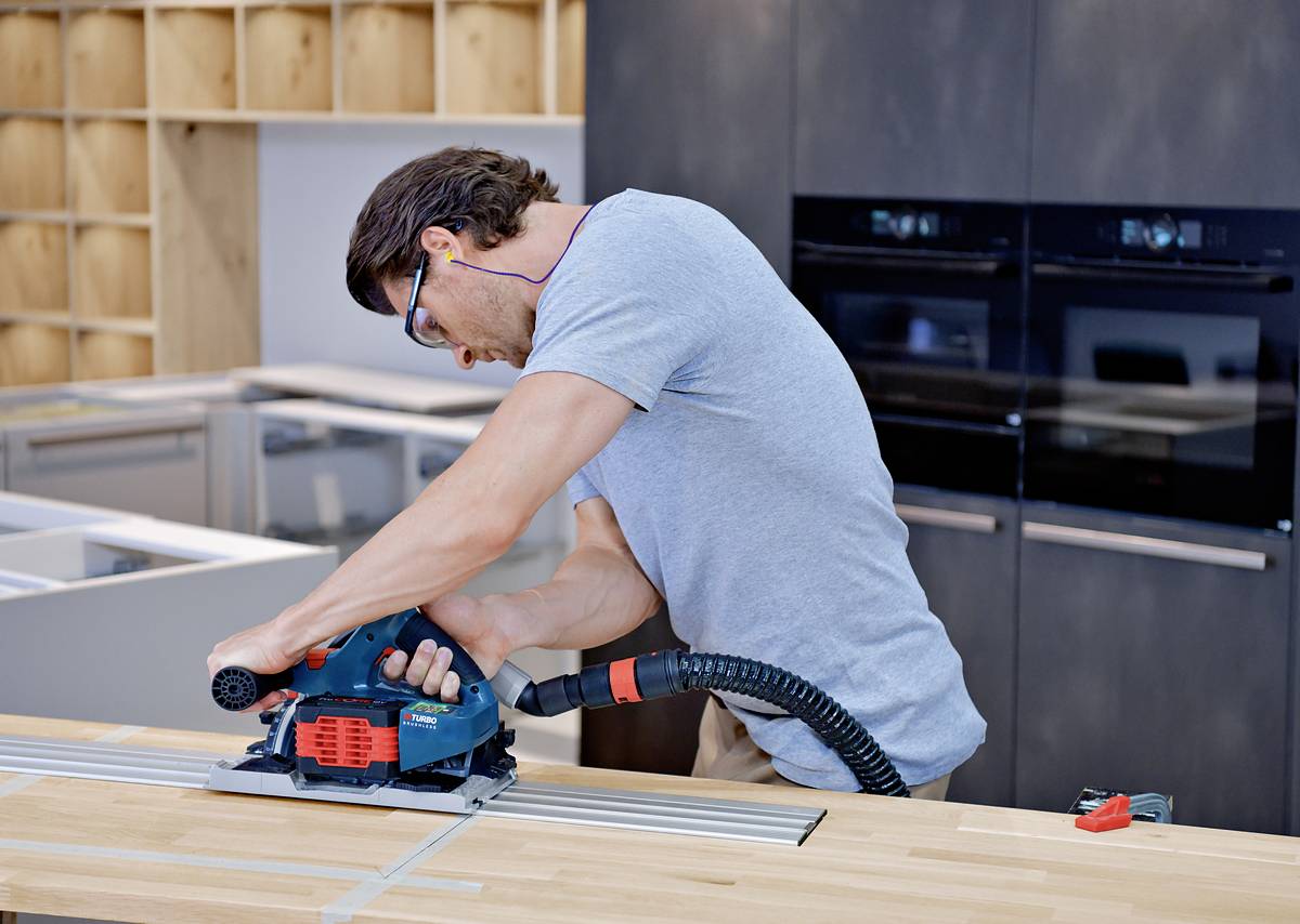 Mężczyzna obrabia blat kuchenny w nowoczesnej kuchni za pomocą elektronarzędzia. Ma na sobie okulary ochronne i zatyczki do uszu.