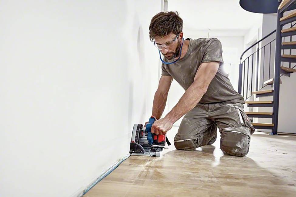 Mężczyzna w ochronnych okularach klęczy na podłodze i za pomocą elektrycznej piły przecina krawędź drewnianej podłogi.