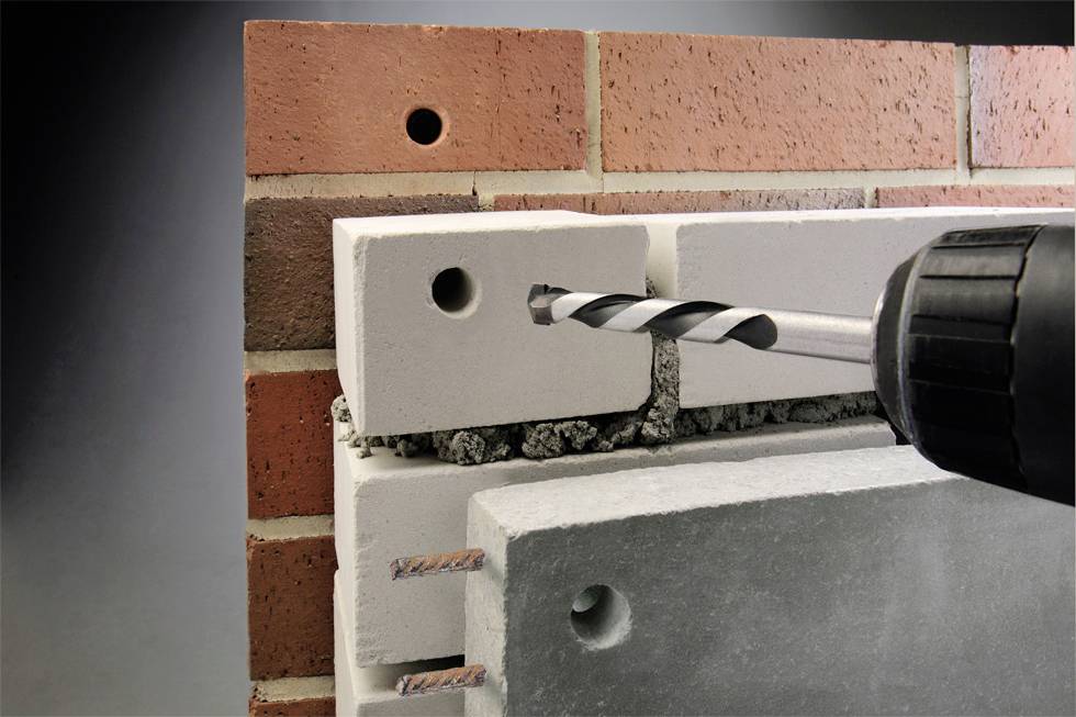 Wiertło jest wprowadzane w płytę betonową z elewacją klinkierową, aby wywiercić otwór. Scena przedstawia proces wiercenia w szczegółach.