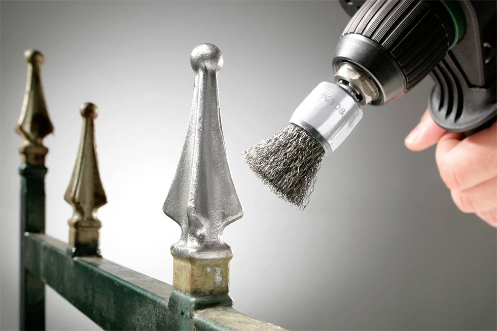 Jedna ręka trzyma wiertarkę z końcówką szczotki drucianej, polerując słup ogrodzeniowy. Różnica między wypolerowaną a niewypolerowaną powierzchnią jest widoczna.