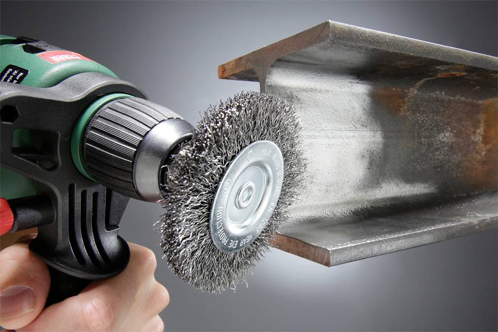 Dłoń trzyma wiertarkę z druciankową szczotką, czyści krawędź metalu. Widoczna powierzchnia nabiera połysku, narzędzie w ruchu.