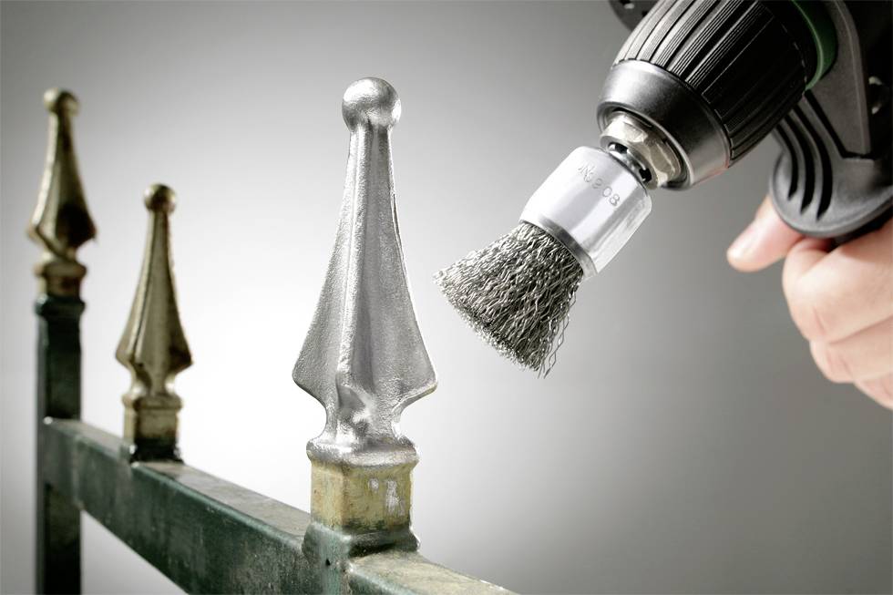 Zbliżenie ogrodzenia polerowanego wiertarką z końcówką w postaci szczotki drucianej. Ostrze metalowego przęsła jest w centrum ostrości.