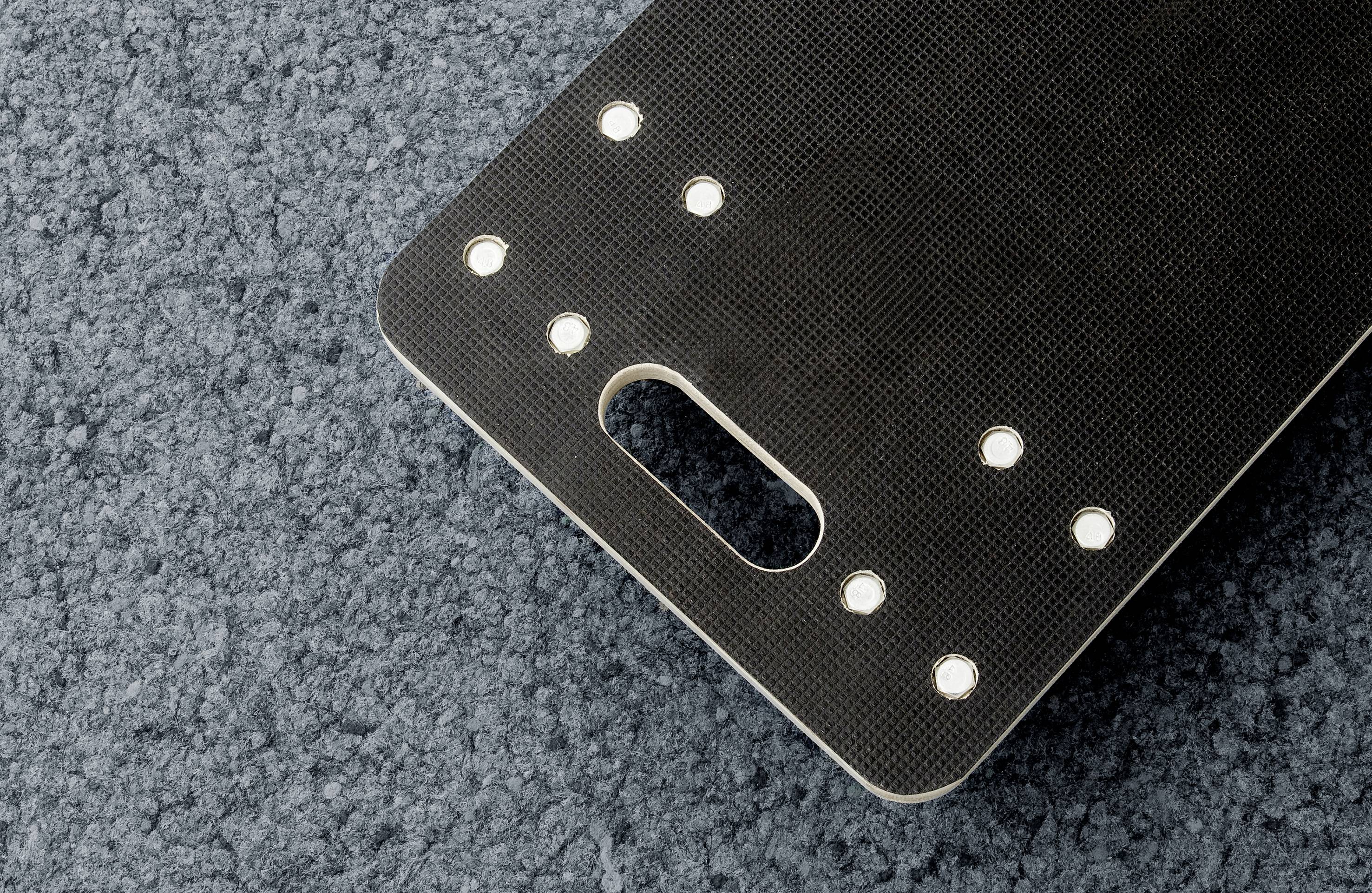 Czarna deska do krojenia z uchwytem i kilkoma okrągłymi światełkami wzdłuż krawędzi leży na szarym dywanie.