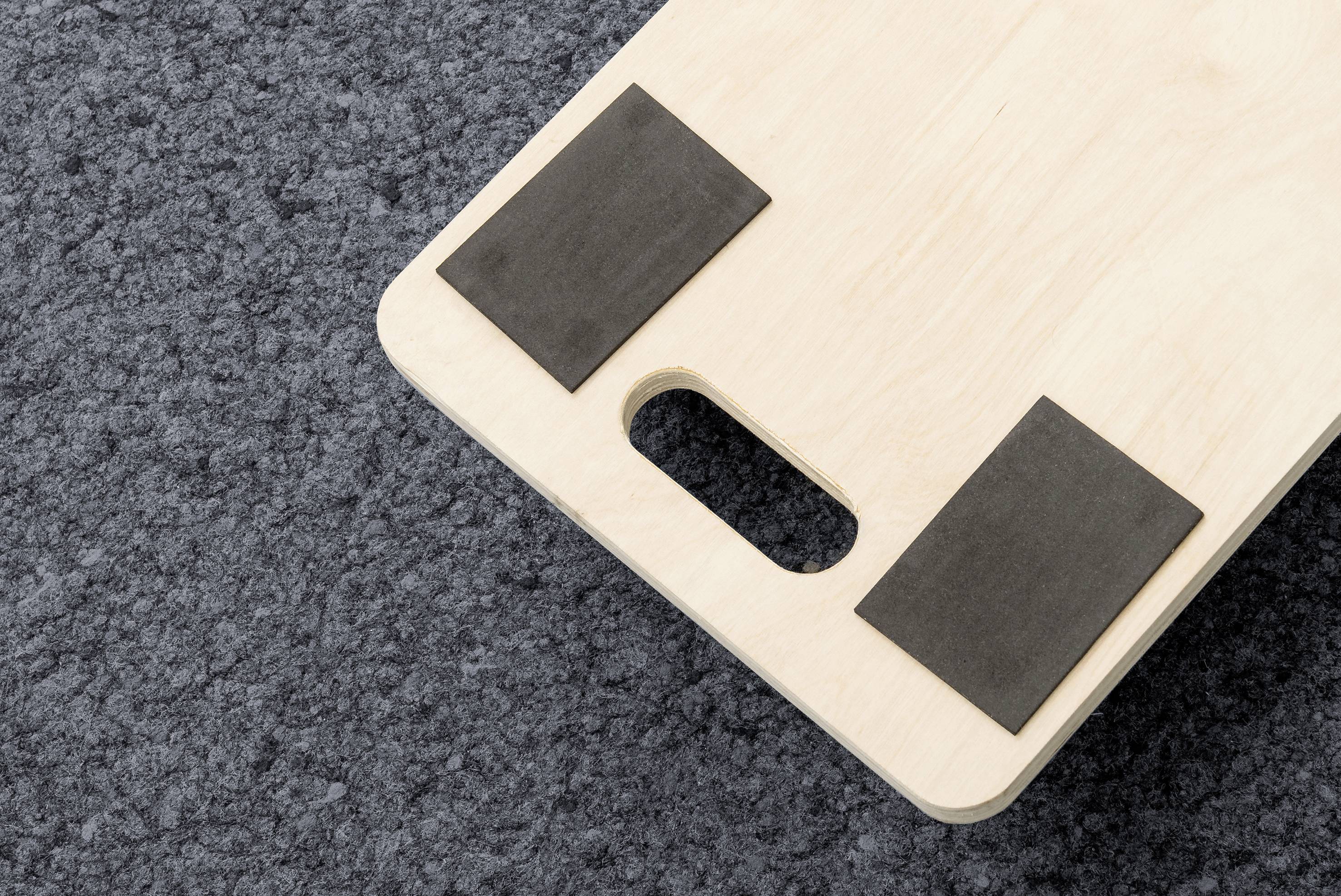 Drewniana deska z dwoma czarnymi gumowymi podkładkami i uchwytem do przenoszenia leży na szarym dywanie.