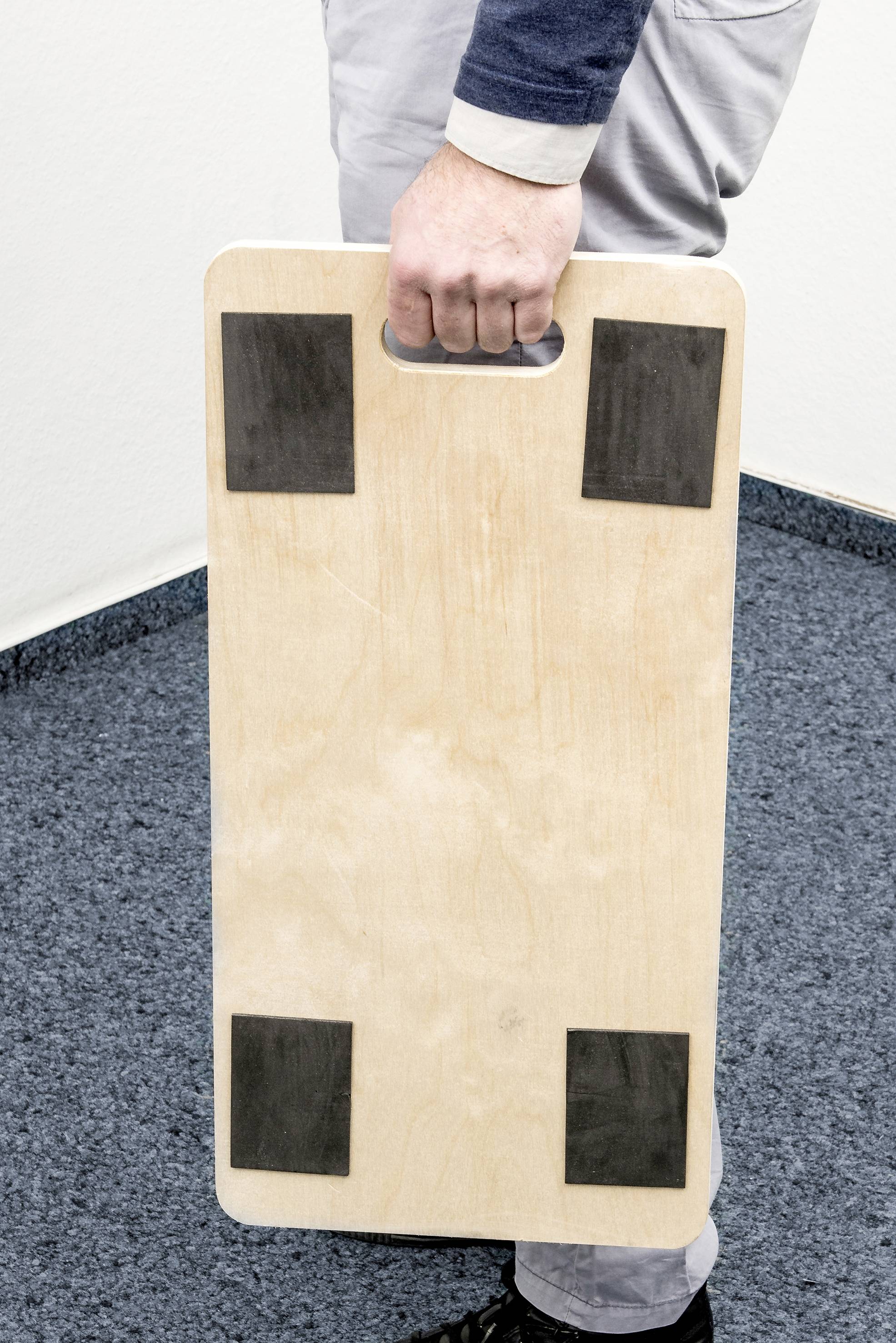 Osoba trzyma prostokątną drewnianą deskę z otworem na chwyt i czterema czarnymi gumowymi podkładkami w rogach, stojąc na niebieskim dywanie.