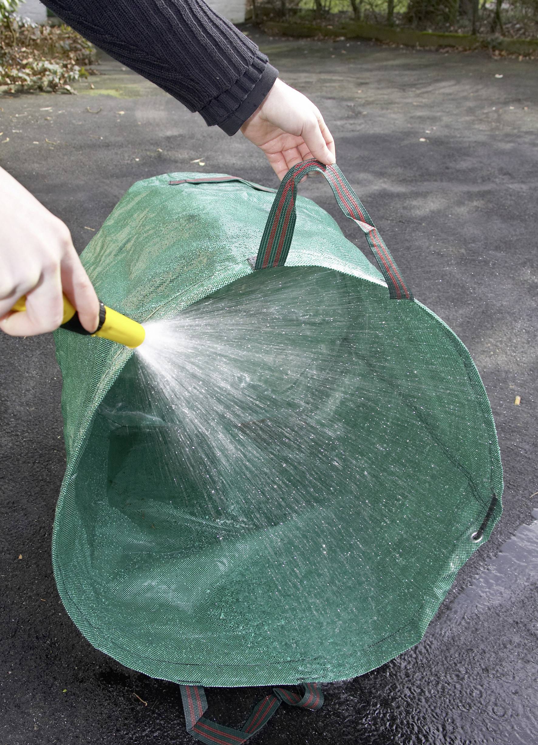 Osoba podlewa wodą z wężem ogrodowym dużą, zieloną, pustą torbę na wolnym powietrzu na asfaltowej powierzchni.