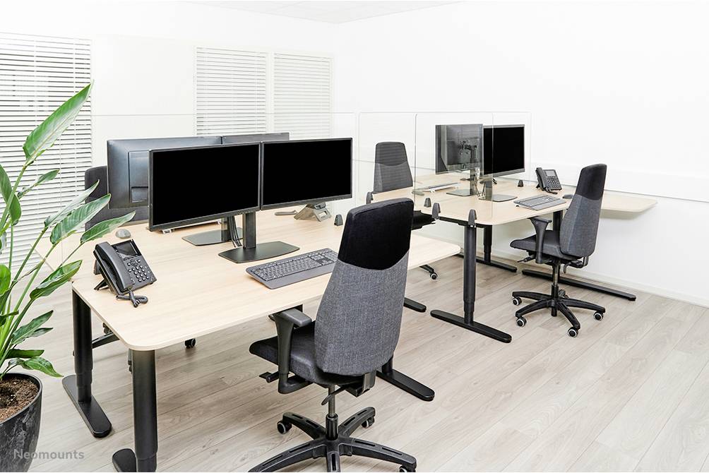 Nowoczesne środowisko biurowe z czterema stanowiskami pracy. Każde stanowisko wyposażone jest w podwójne monitory, ergonomiczny fotel biurowy i telefon. Na pierwszym planie znajduje się zielona roślina.