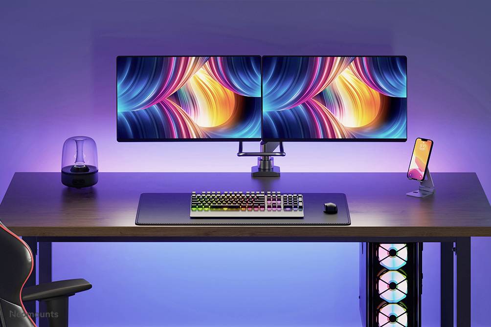 Biurko z dwoma jaskrawo kolorowymi monitorami, klawiaturą RGB, smartfonem i lampką, otoczone niebieskim podświetleniem w tle.