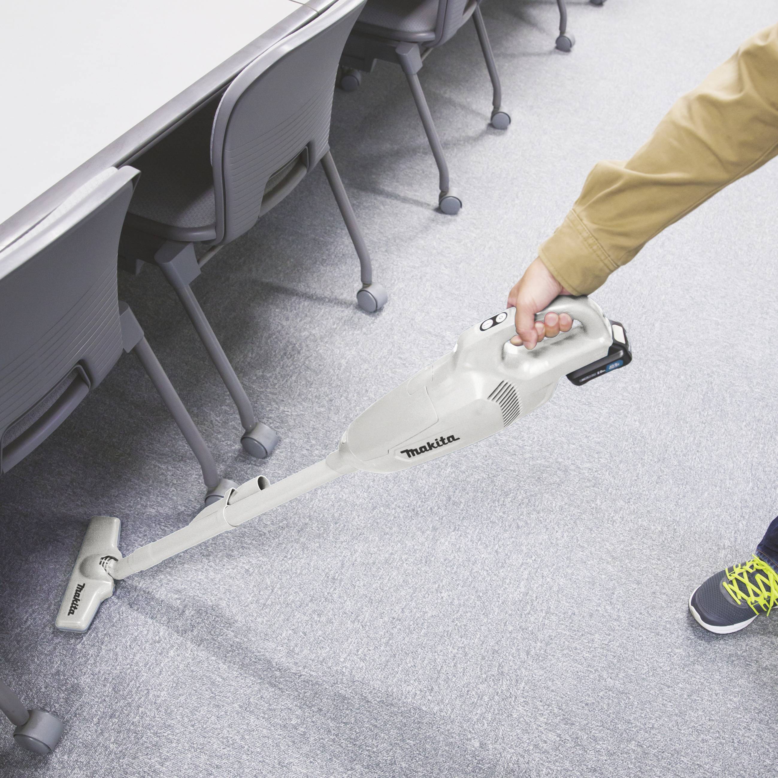Osoba odkurza podłogę dywanową przy stole za pomocą bezprzewodowego odkurzacza. W pobliżu stoi rząd krzeseł, ukazując proces sprzątania.