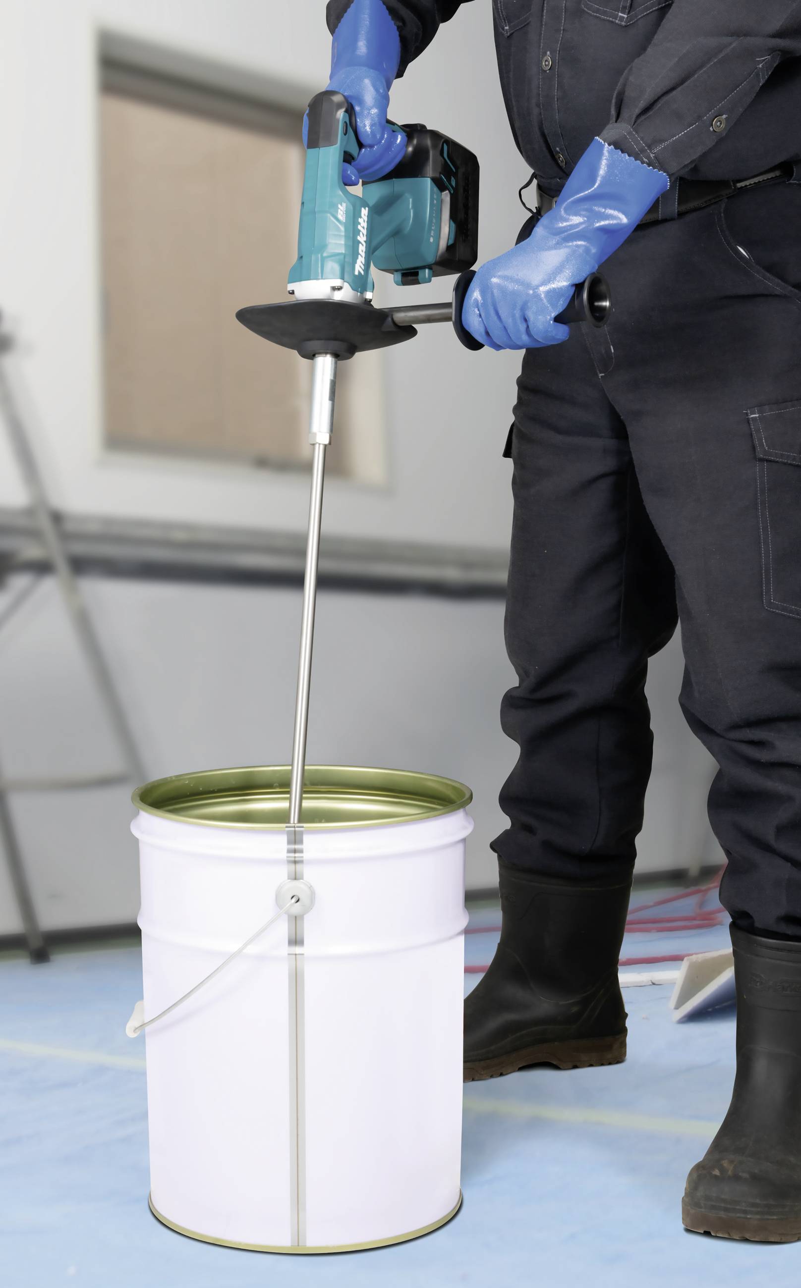 Osoba miesza farbę elektrycznym mieszadłem w dużym białym wiadrze na niebieskiej podłodze.