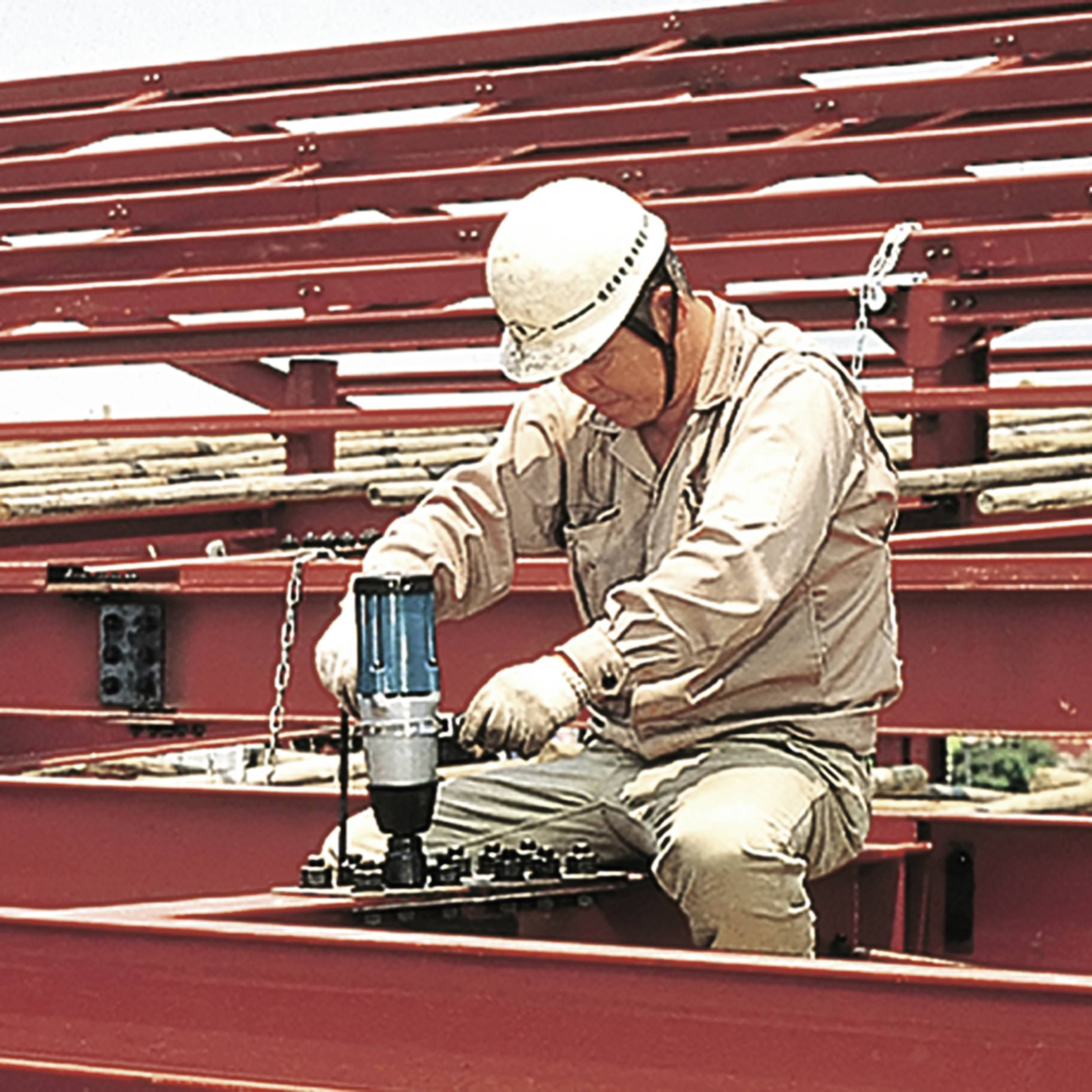 Robotnik budowlany w odzieży ochronnej i kasku używa elektronarzędzia do obróbki stalowych belek na placu budowy.