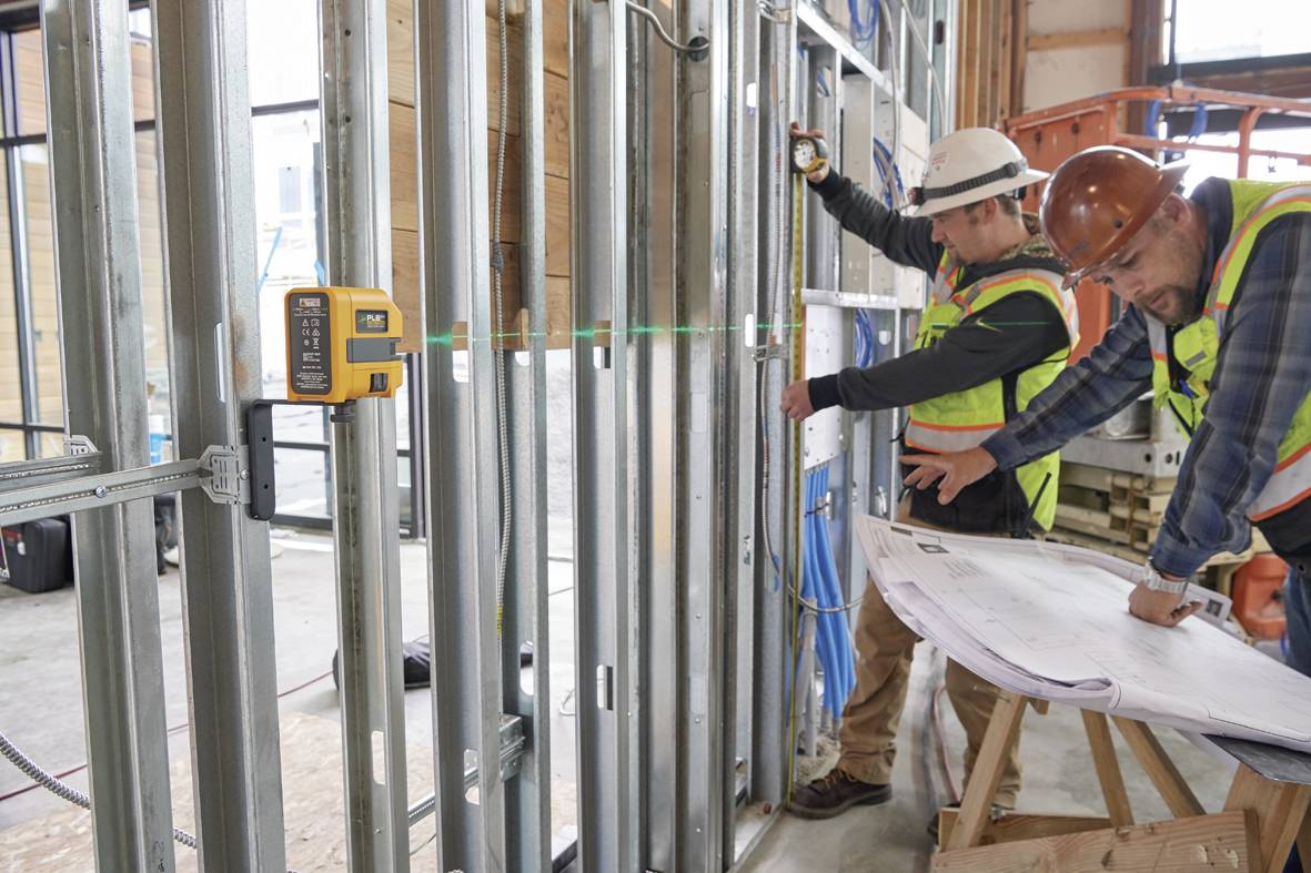 Robotnicy budowlani na placu budowy sprawdzają plany budowlane przed metalową konstrukcją szkieletową. Na ścianie zamocowane jest urządzenie do laserowego poziomowania.