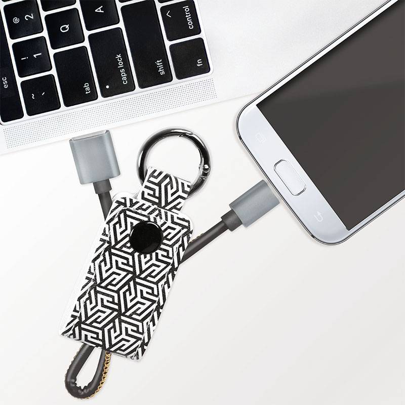 Smartfon leży obok klawiatury laptopa. Widoczny jest kabel ładujący oraz brelok do kluczy z geometrycznym wzorem.