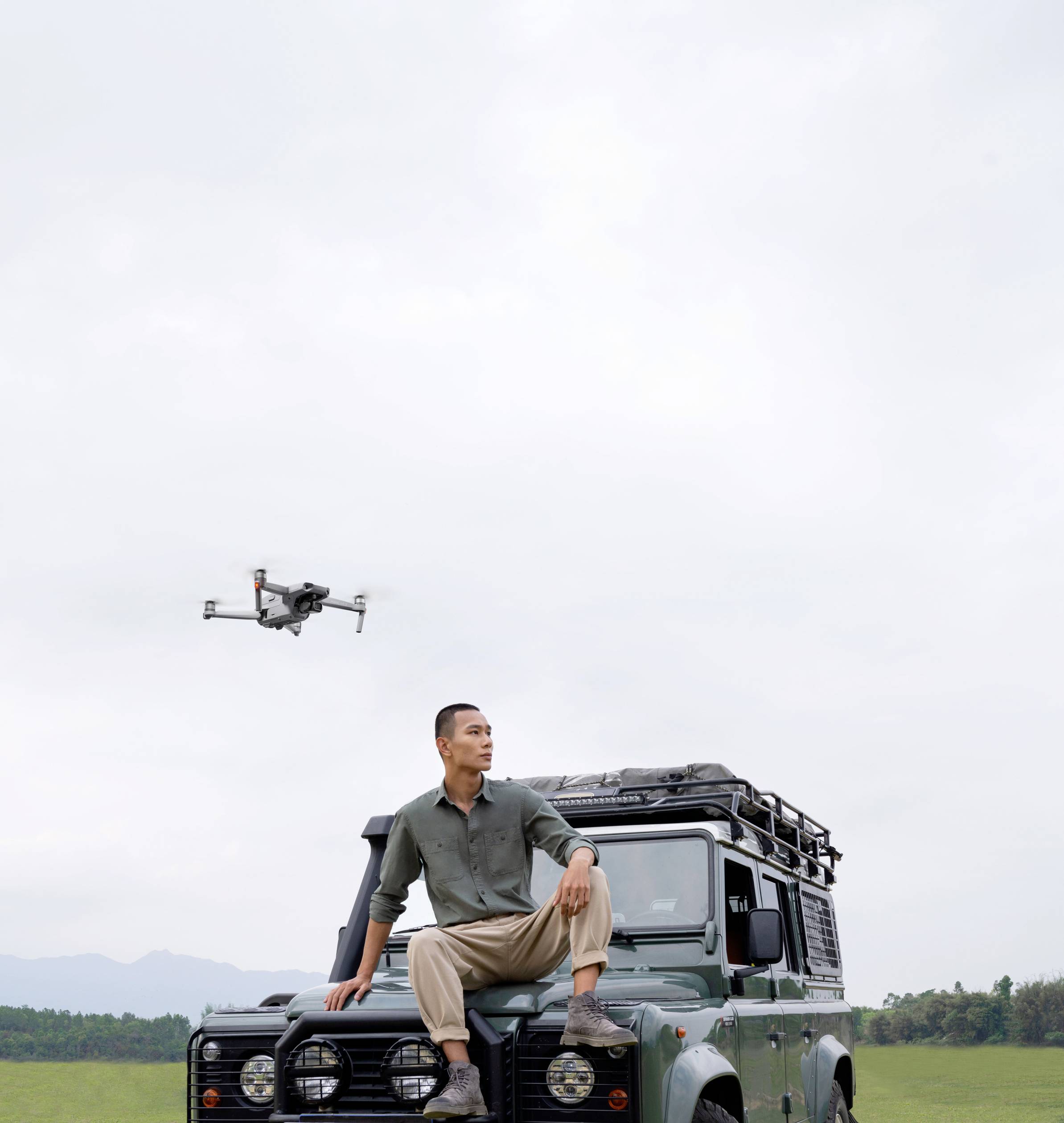 Mężczyzna siedzi na zielonym samochodzie terenowym na polu, podczas gdy dron unosi się w powietrzu. Niebo jest zachmurzone.