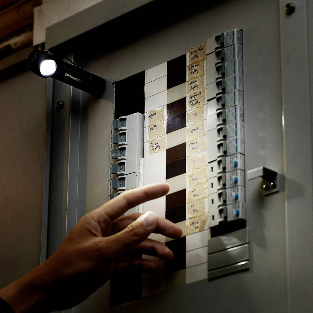 Jedna ręka wskazuje na otwarty bezpiecznikowy skrzynkę z wieloma przełącznikami. Latarka oświetla skrzynkę.