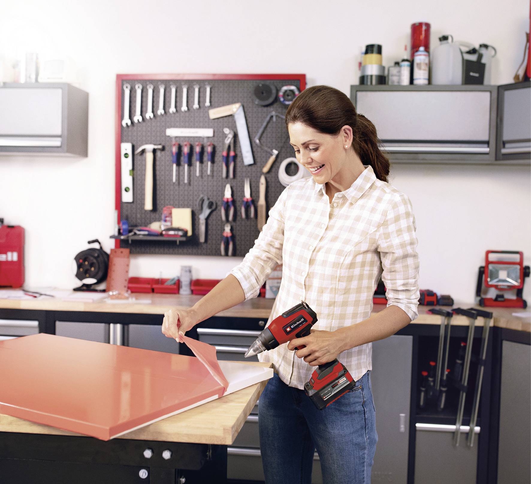 Osoba w warsztacie pracuje nad pomarańczową blachą za pomocą akumulatorowego wkrętaka. W tle wiszą różne narzędzia.