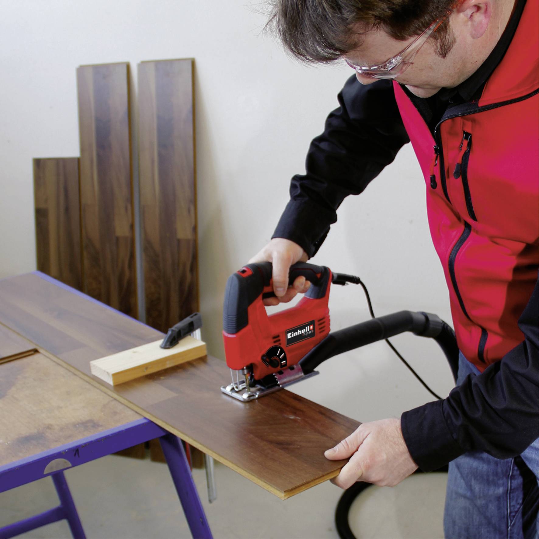 Osoba w czerwonej kurtce roboczej przecina drewnianą deskę czerwoną piłą elektryczną na stole warsztatowym.