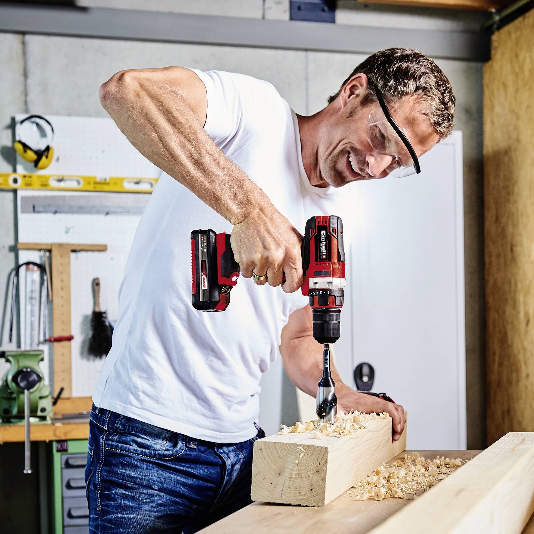 Mężczyzna w okularach ochronnych i białej koszulce wierci dziurę akumulatorowym wiertłem w desce w środowisku warsztatu.