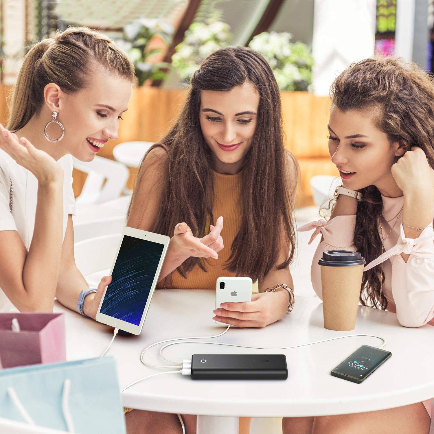 Trzy młode kobiety siedzą przy stole, korzystając ze smartfonów i tabletu podłączonego do powerbanku. Śmieją się i wyglądają na bardzo rozbawione.