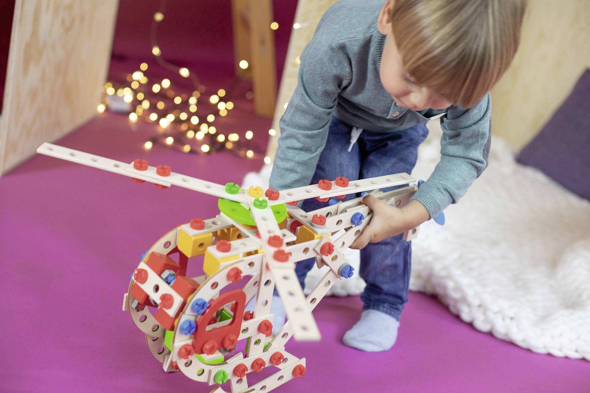 Dziecko bawi się kolorowym zestawem klocków w kształcie helikoptera na różowym dywanie, otoczone girlandami świetlnymi.
